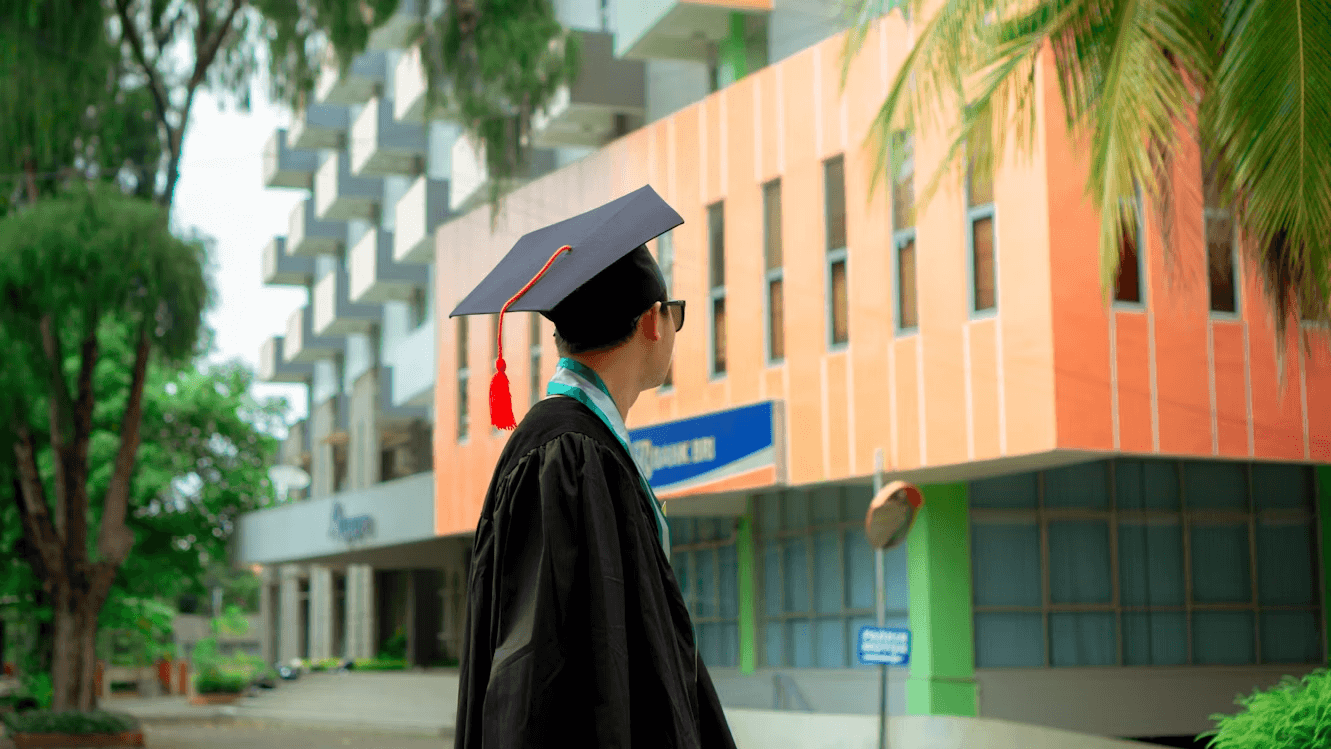 Graduate in a black cap and gown standing on a university campus looking toward modern academic buildings