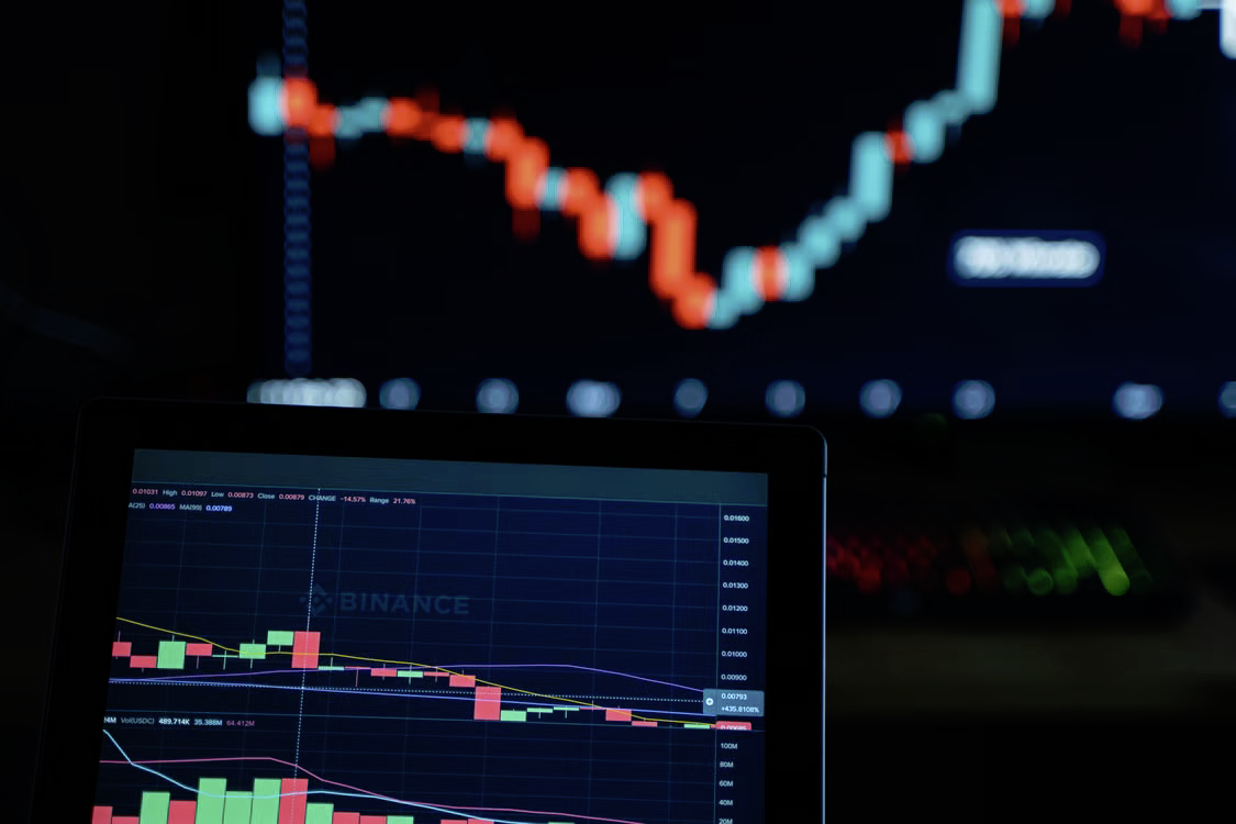 Tablet displaying a Binance cryptocurrency candlestick trading chart with stock market graphs on a monitor in the background