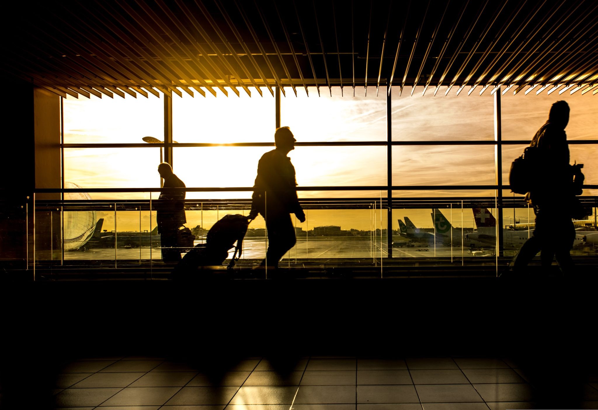 Silhouettes of travelers pulling luggage through an airport terminal corridor with airplanes parked on the tarmac at sunset