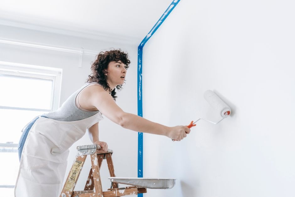 Painter on a step ladder rolling white paint onto an interior wall lined with blue painter's tape